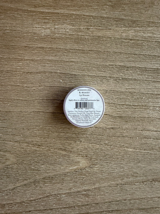 A small round tin of Morphe Cosmetics lip gloss, showing the white label with a barcode, placed on a light brown wood grain surface.