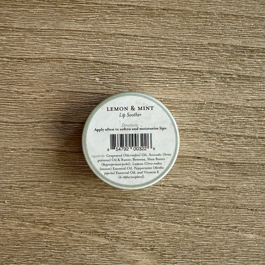 A small round metal tin of lip balm or lip soother labeled 'LEMON & MINT' with a barcode, placed on a light brown wood-grain surface.