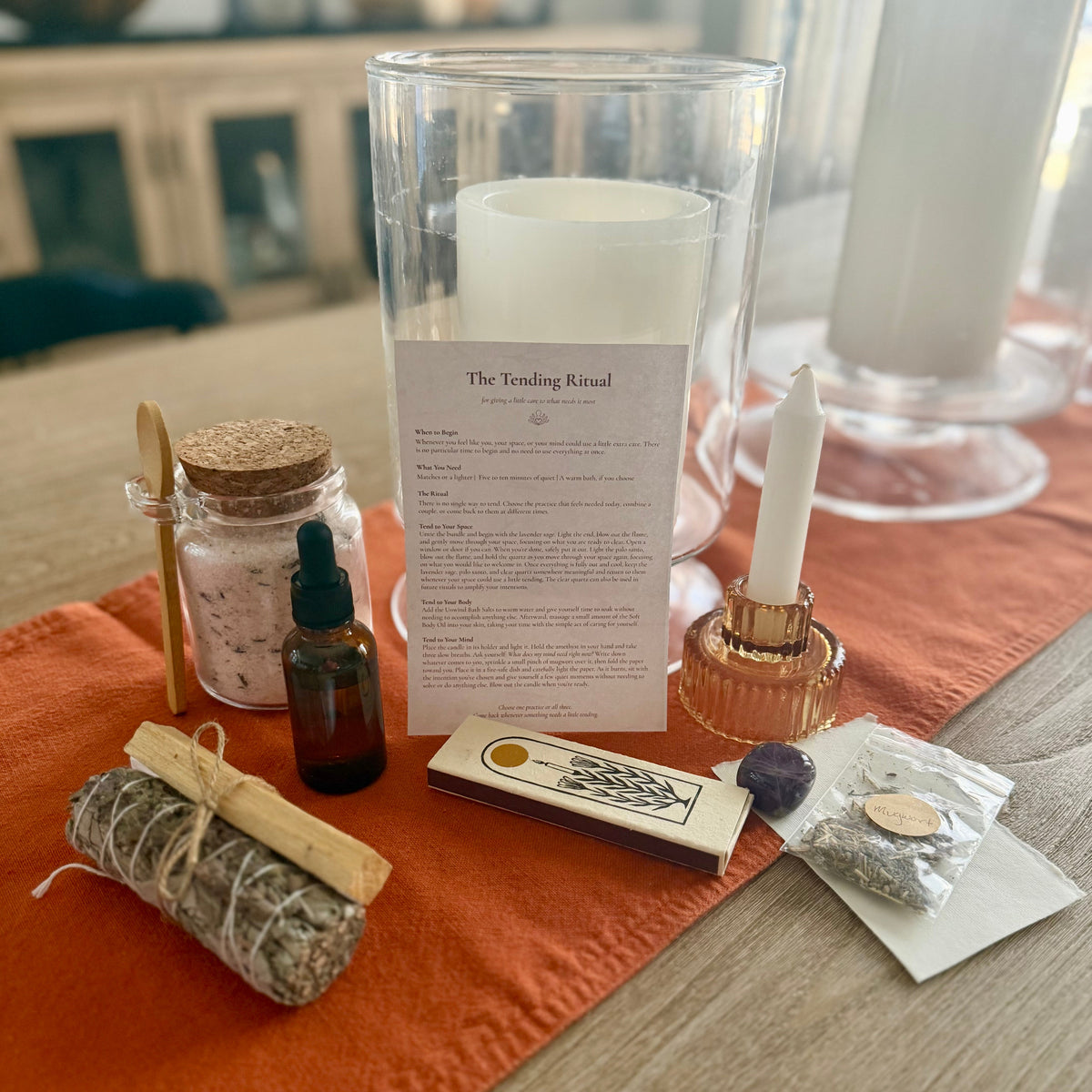 A ritual kit arranged on an orange runner on a wooden table, featuring a white candle in a clear glass holder, a small amber dropper bottle, a sage bundle tied with string, a matchbox, a small pouch, and a printed instruction card titled 'The Tending Ritual'.