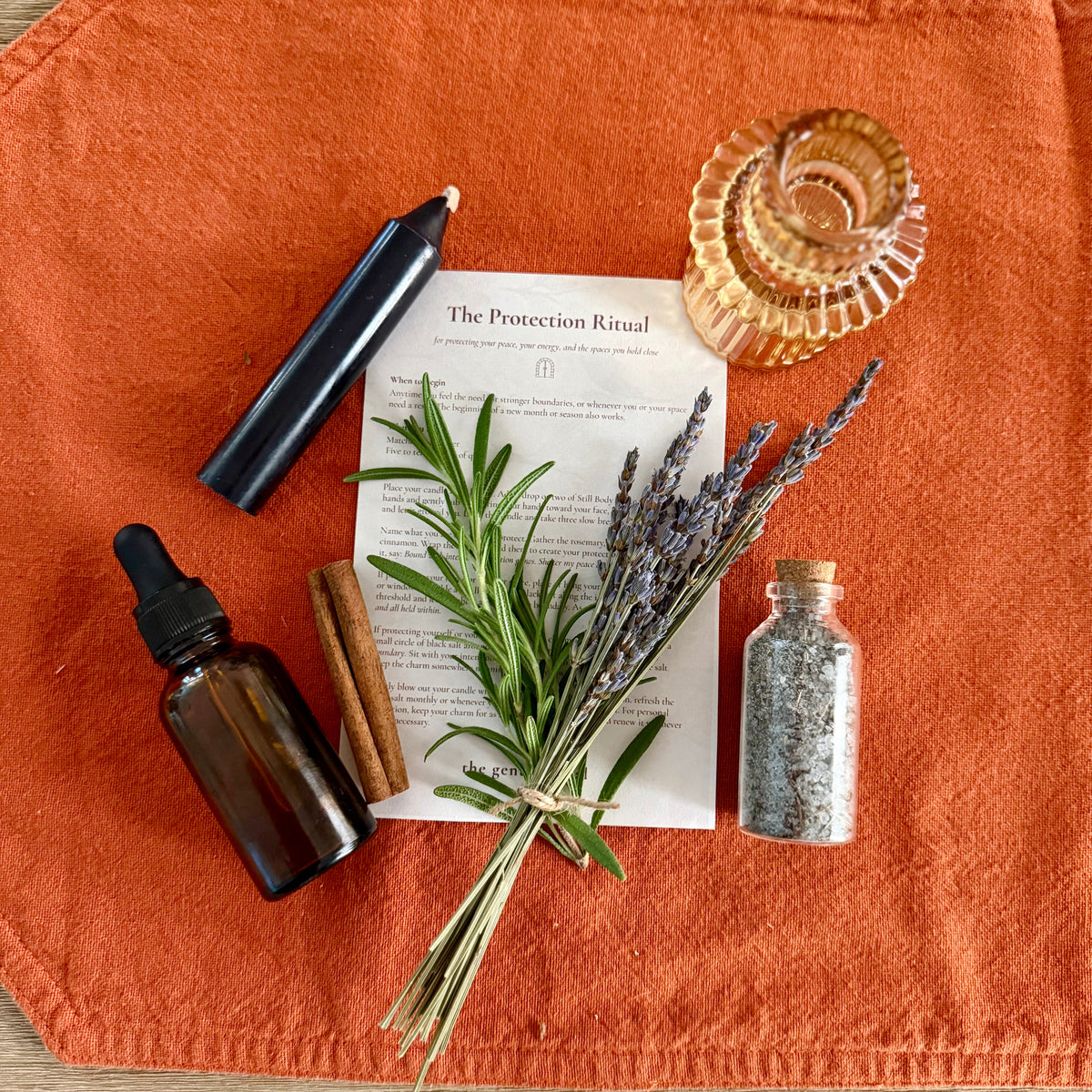 An autumn-themed ritual setup featuring a bundle of lavender, cinnamon sticks, a small glass bottle of salt, a black candle, and an amber dropper bottle, arranged on an orange fabric cloth over a light wood background.