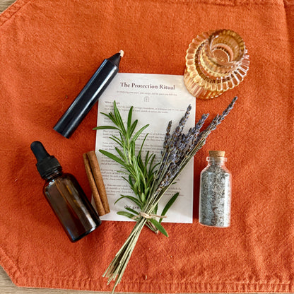 An autumn-themed ritual setup featuring a bundle of lavender, cinnamon sticks, a small glass bottle of salt, a black candle, and an amber dropper bottle, arranged on an orange fabric cloth over a light wood background.