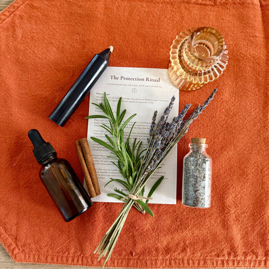 An autumn-themed ritual setup featuring a bundle of lavender, cinnamon sticks, a small glass bottle of salt, a black candle, and an amber dropper bottle, arranged on an orange fabric cloth over a light wood background.