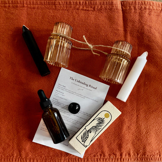 A styled flat-lay of a ritual or candle-making kit on an orange linen cloth over a wood surface. The kit includes two amber glass candle holders tied with twine, a black wand, a white wax stick, an amber dropper bottle, a white candle, a printed instruction sheet titled 'The Unbinding Ritual', a black sphere, and a wooden box with a bird illustration.